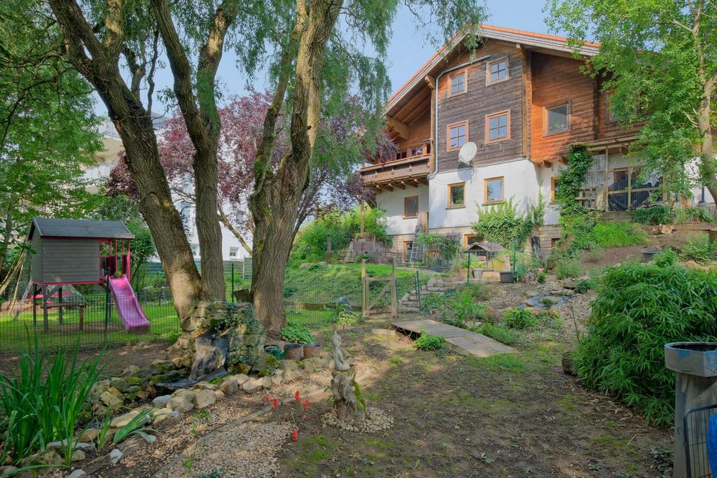 Large wooden and stone house with a garden, slide and playground in the foreground, surrounded by trees and plants.