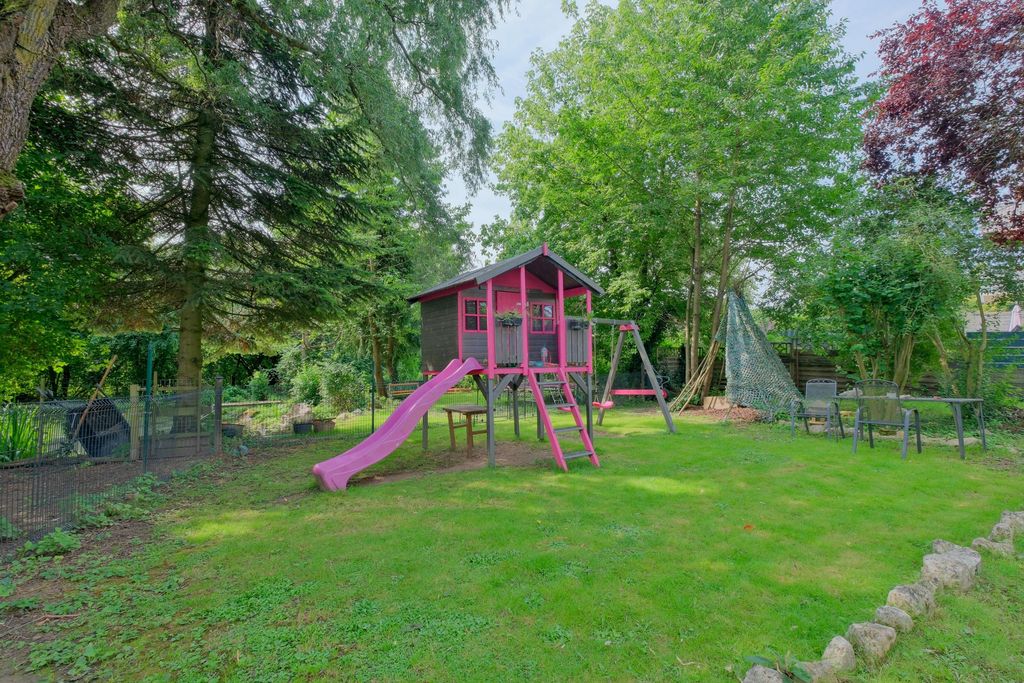 Children's playground in a garden featuring a wooden playhouse with a pink slide, swing set, and climbing net, surrounded by trees and grassy areas.