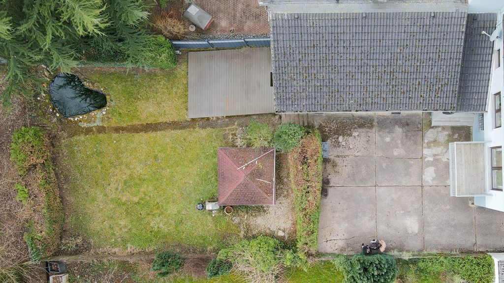 Drone view of a small garden with a gazebo in the center, a pond on the left side, and a house with a chimney on the right.