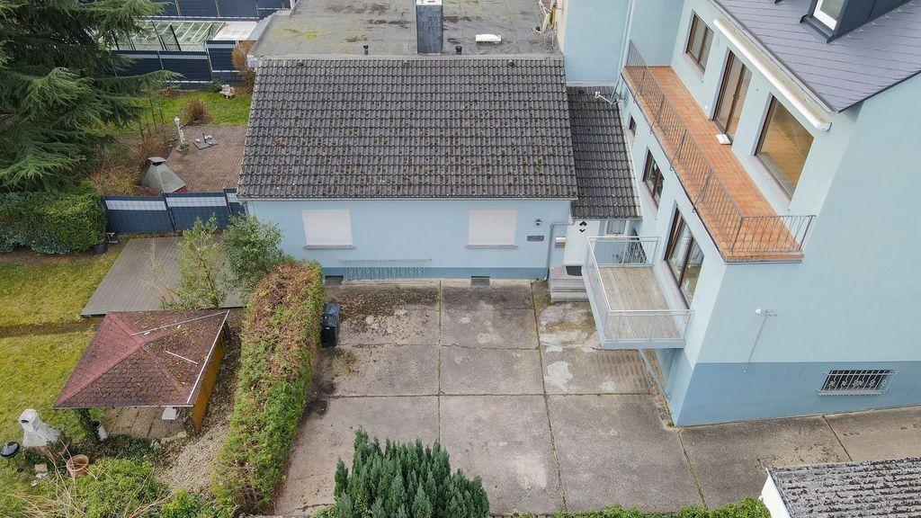 Bird's-eye view of a blue-gray residential house with a balcony and adjacent garage, surrounded by a green garden and paved courtyard.