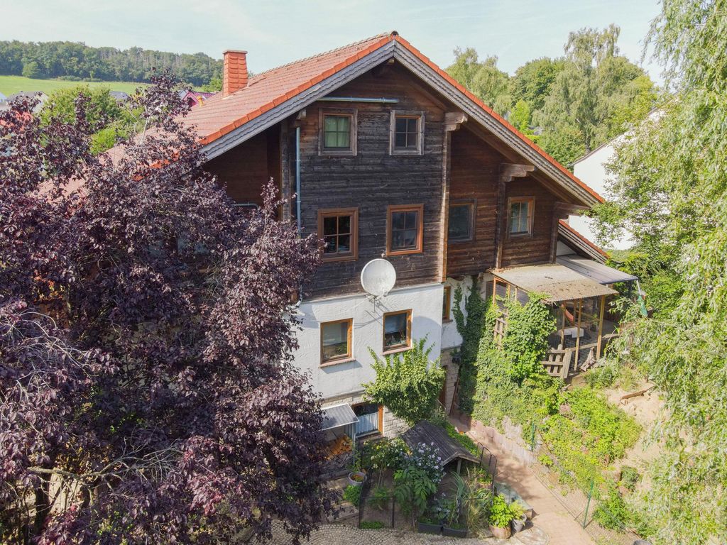 A traditional multi-story wooden house with a red tiled roof, surrounded by green trees and plants in a rural setting.