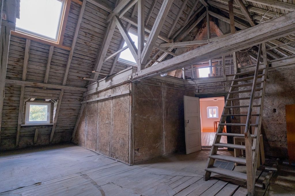 Old, empty attic with visible wooden beams, a small staircase, and an open door leading to a bright wall with a window on the opposite side.