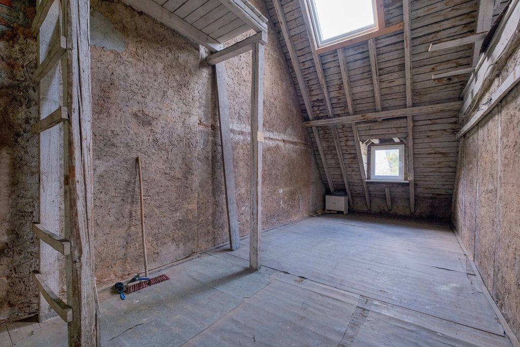 An empty old attic with slanted wooden walls and a window in the gable roof, featuring visible wooden beams and a broom leaning against the wall.