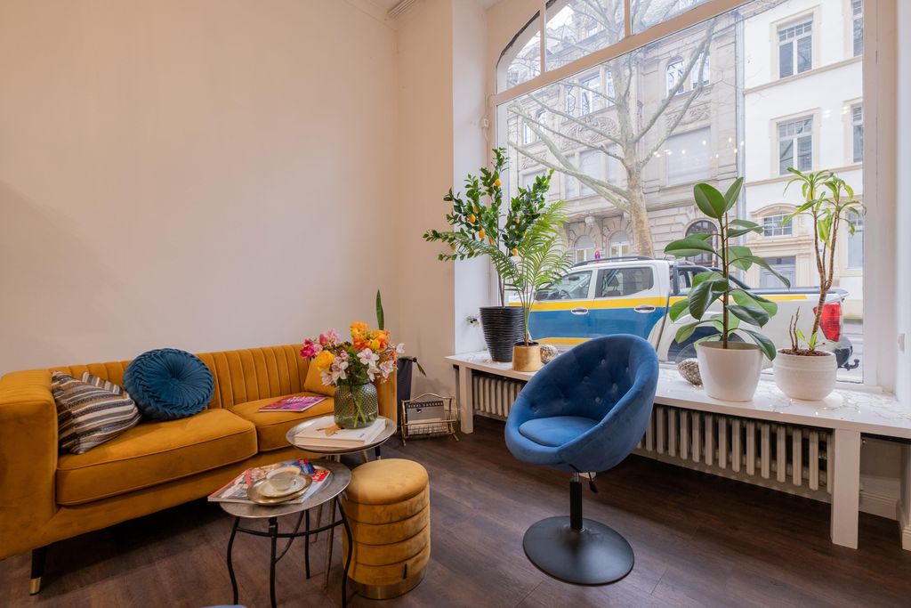 Cozy living room with an orange velvet sofa, a blue velvet chair, and several plants on the windowsill of a large window overlooking a street.
