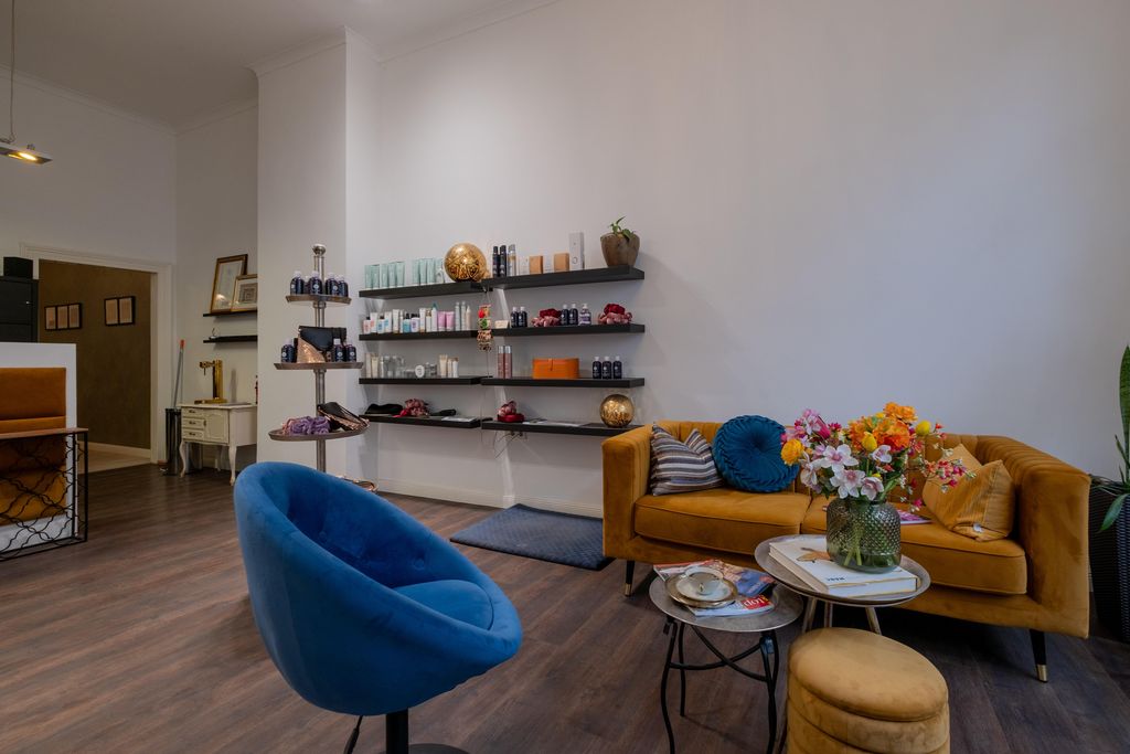 A cozy waiting area featuring a blue armchair, an orange sofa, a small coffee table with magazines, and a vase of colorful flowers. Shelves with various cosmetic and skincare products are mounted on the wall.