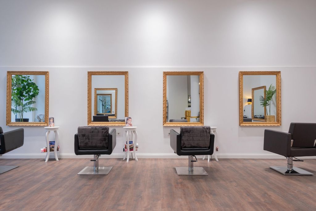 Interior of a modern hair salon with four styling chairs in front of large wall mirrors, small white tables in between, and wooden flooring.