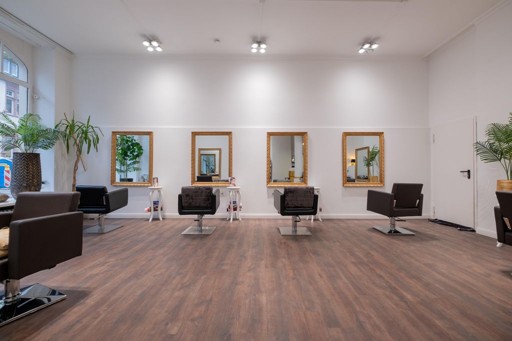 Modern hair salon with multiple styling chairs in front of large mirrors, wooden floor, and plants along the walls.