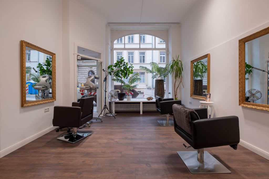 Interior of a hair salon with two barber chairs, large mirrors on the walls, and green plants in front of a large window.