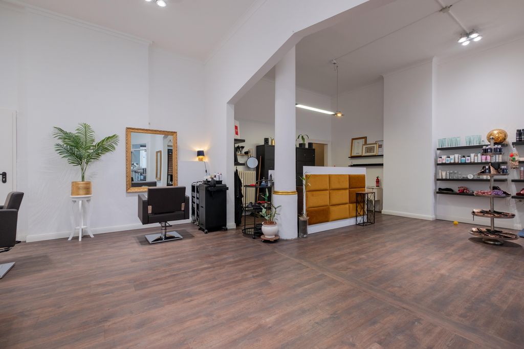 An empty hair salon with two styling chairs, a large mirror, a plant in a golden pot, and a reception desk with brown upholstery.