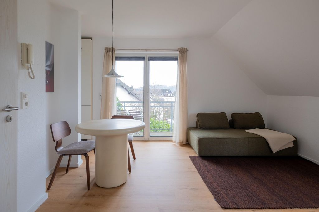 Bright room with a round white dining table, two chairs, and a green sofa with cushions and a blanket next to a large window overlooking houses and trees.