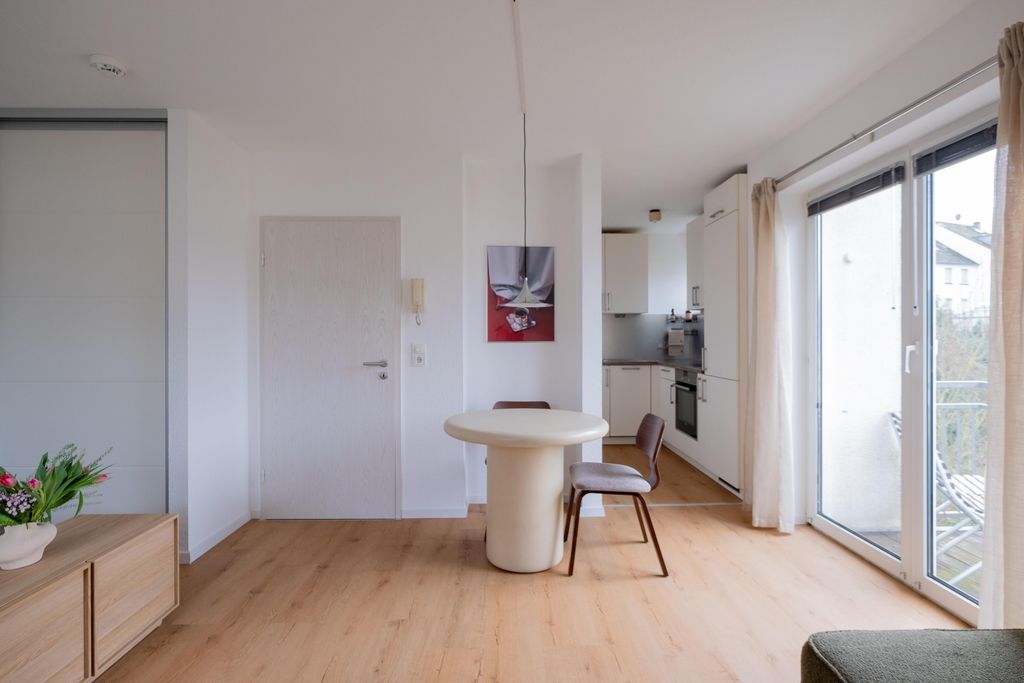 A modern, bright dining area with a round white table and two brown chairs in front of a white door, with a kitchen in the background and large windows leading to a balcony.