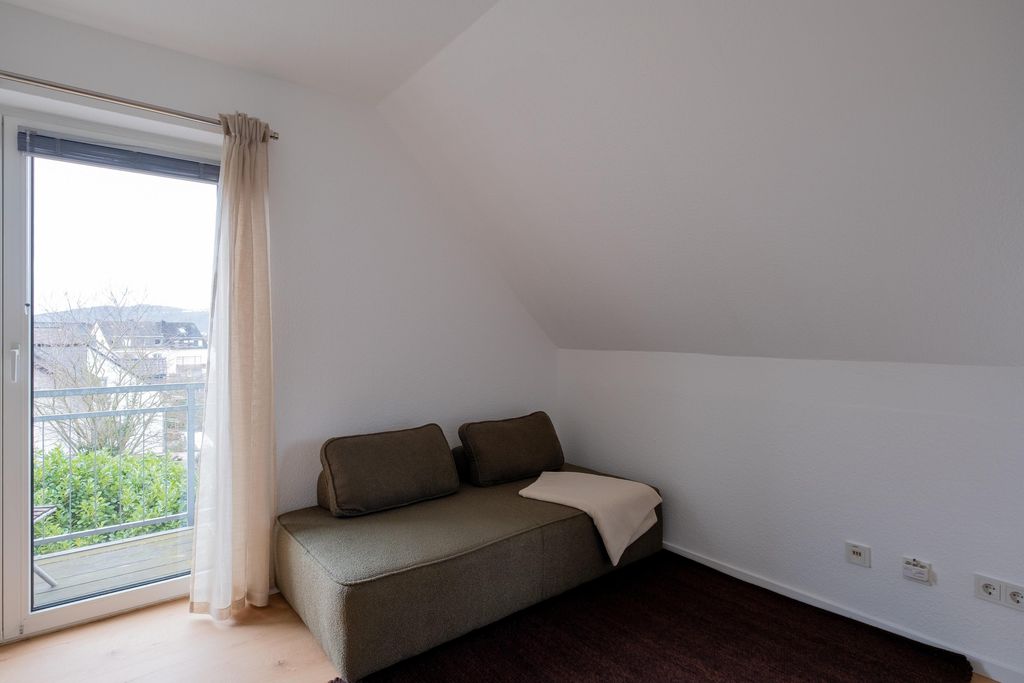 A simple room with a gray sofa, two back cushions, and a light throw next to a balcony door with semi-transparent curtains.