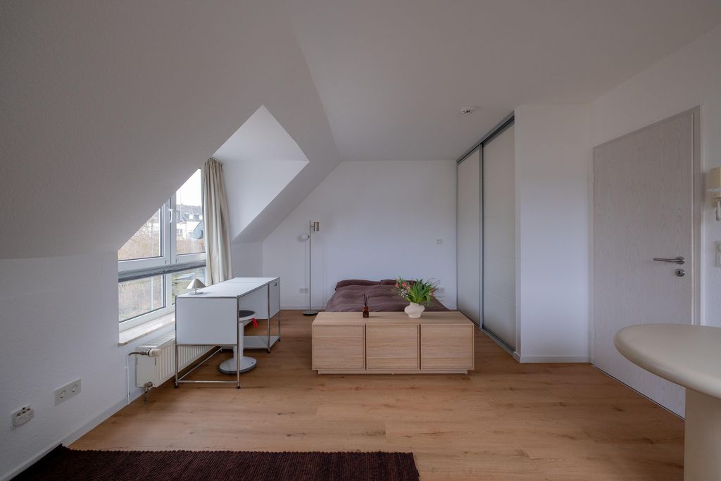 Attic bedroom with large window, desk, double bed with brown duvet and wooden cabinet with plant vases.