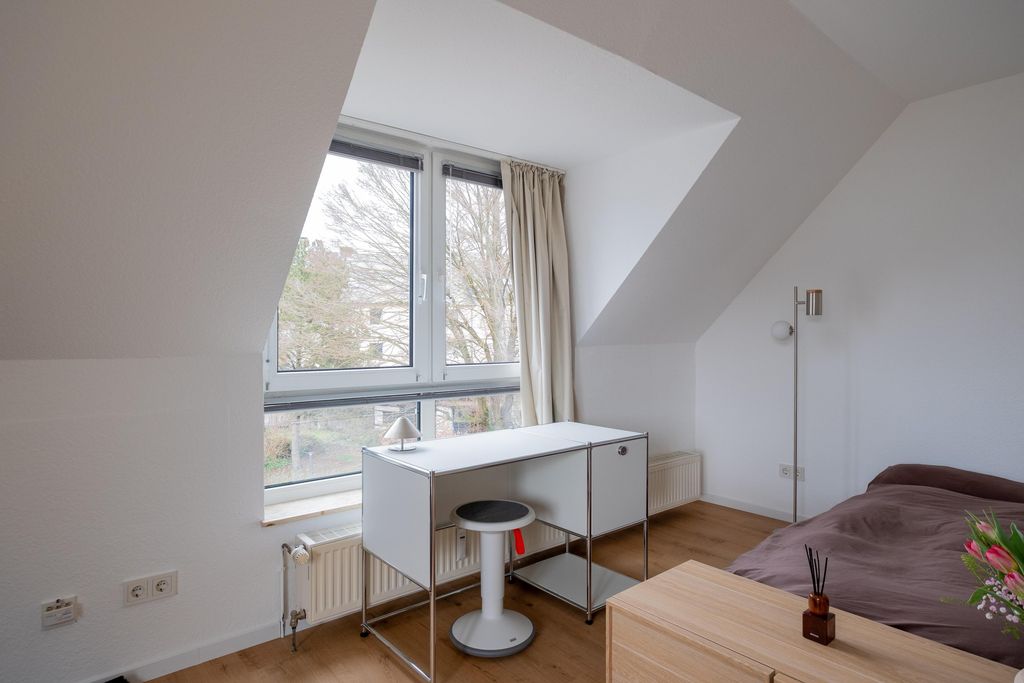 Bright bedroom with slanted ceiling, large window, white desk with stool, floor lamp, and a bed with brown cover.