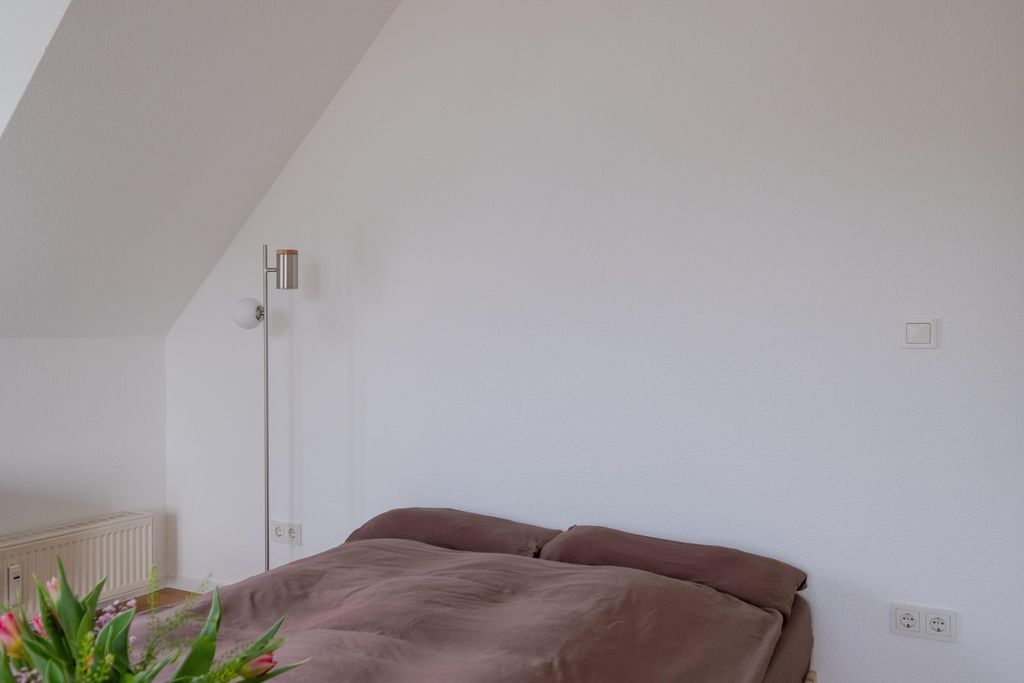 A simple bedroom with a large bed featuring brown pillows against a white wall. Plant leaves are visible in the foreground.