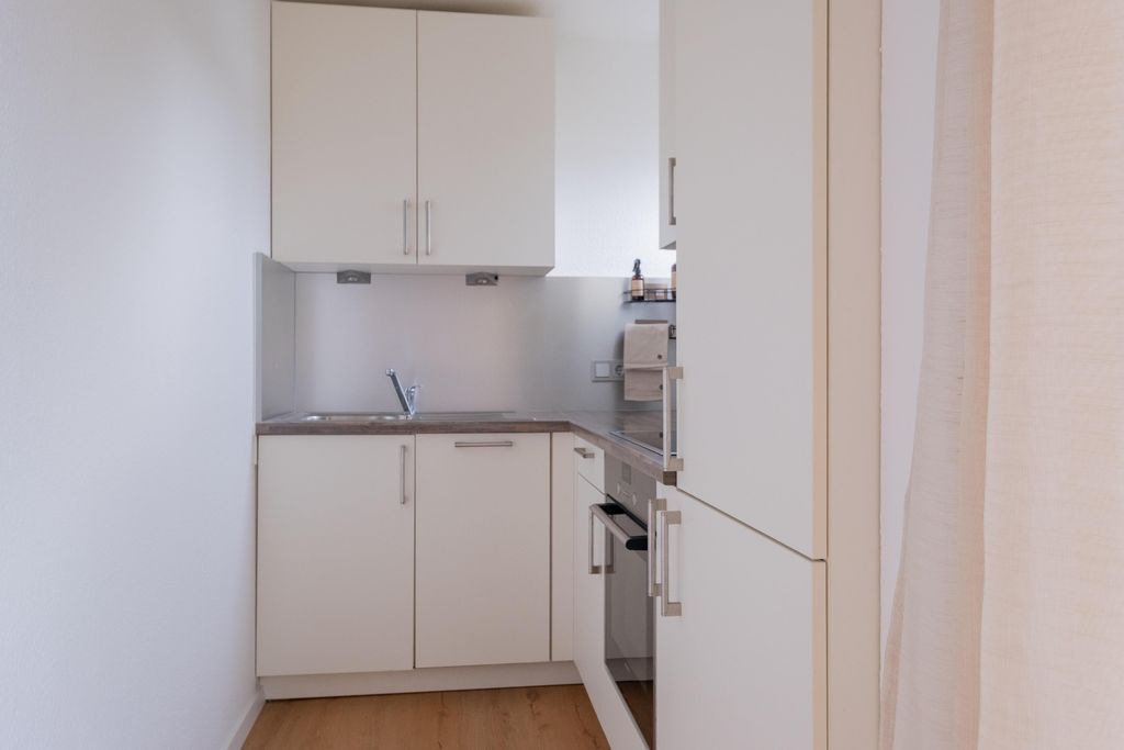 Small modern kitchen with white lacquered cabinets, sink, oven, and wooden flooring.