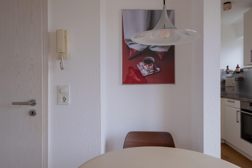 A round dining table with a wooden chair in front of a white wall featuring a modern hanging lamp and a framed picture of a coffee cup on a tray.
