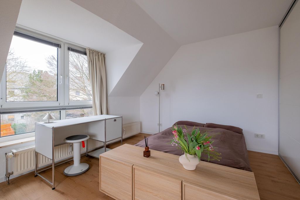 A bright bedroom with a large window, a simple desk with a stool, a double bed with brown bedding, and a wooden dresser with a vase of flowers on top.