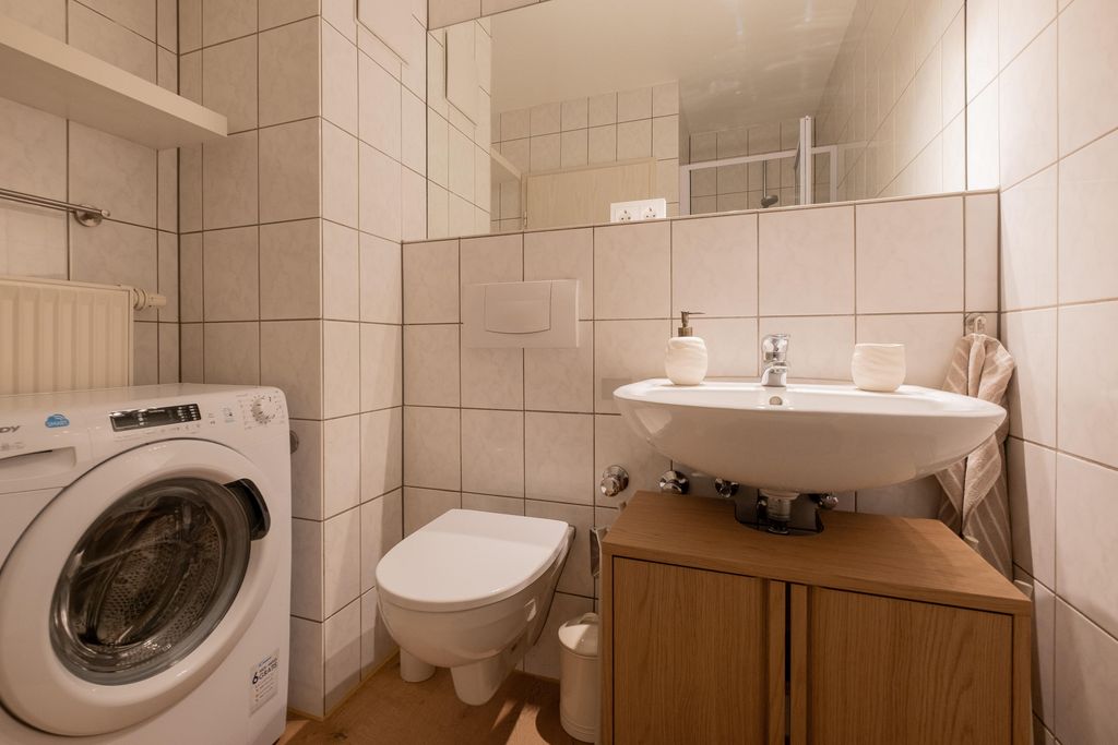 Small bathroom with washing machine, wall-mounted toilet, sink on wooden cabinet, and large mirror.