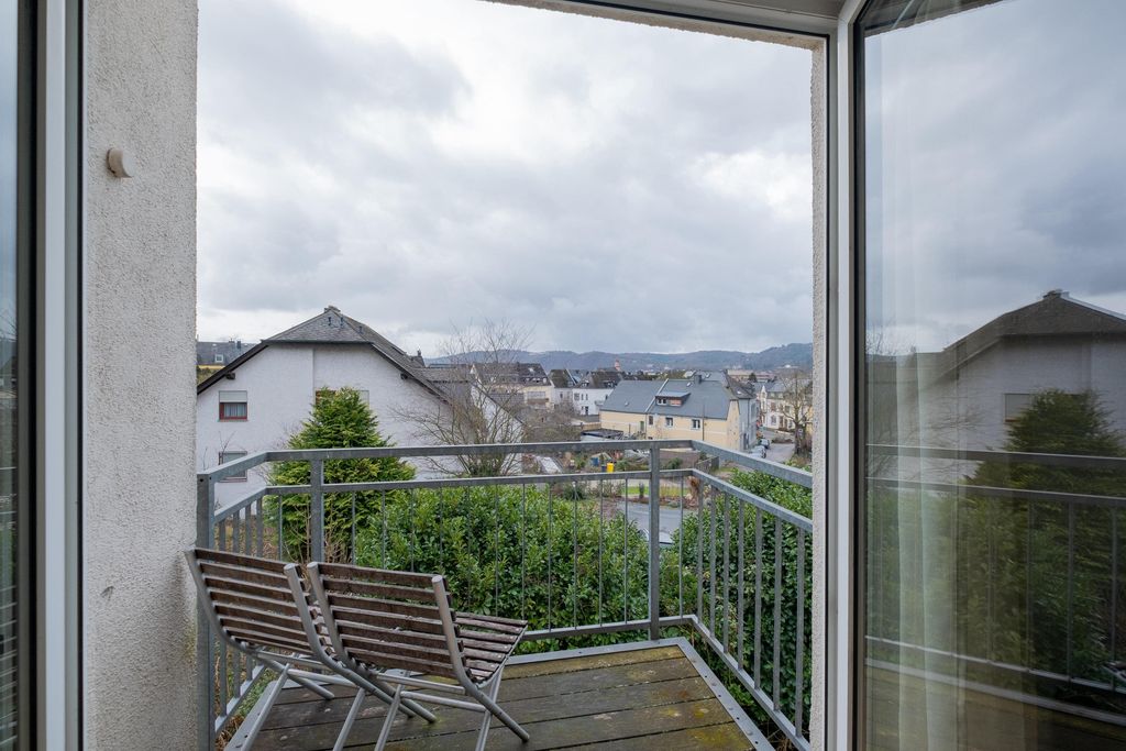 Balcony with two wooden chairs and railing, overlooking a leafy residential area under a cloudy sky.