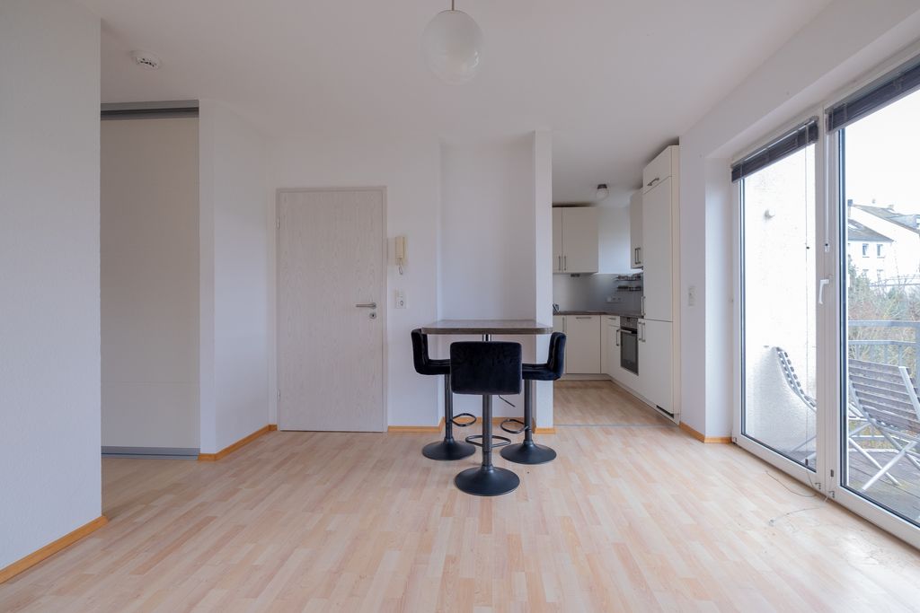 Modern, empty living space with light wood laminate flooring, a small bar with three black bar stools in the center, and an open kitchen in the background. Large windows overlooking a balcony and bright walls with a closed door.