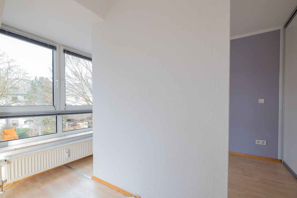 Empty room with light wooden floors and large windows letting in daylight, showing trees and residential houses outside.