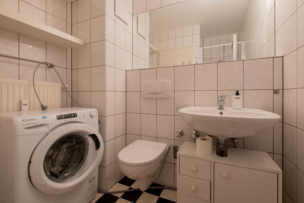A small bathroom featuring a washing machine, a wall-mounted toilet, and a sink on a cabinet, with white tiled walls and a black-and-white checkered floor.
