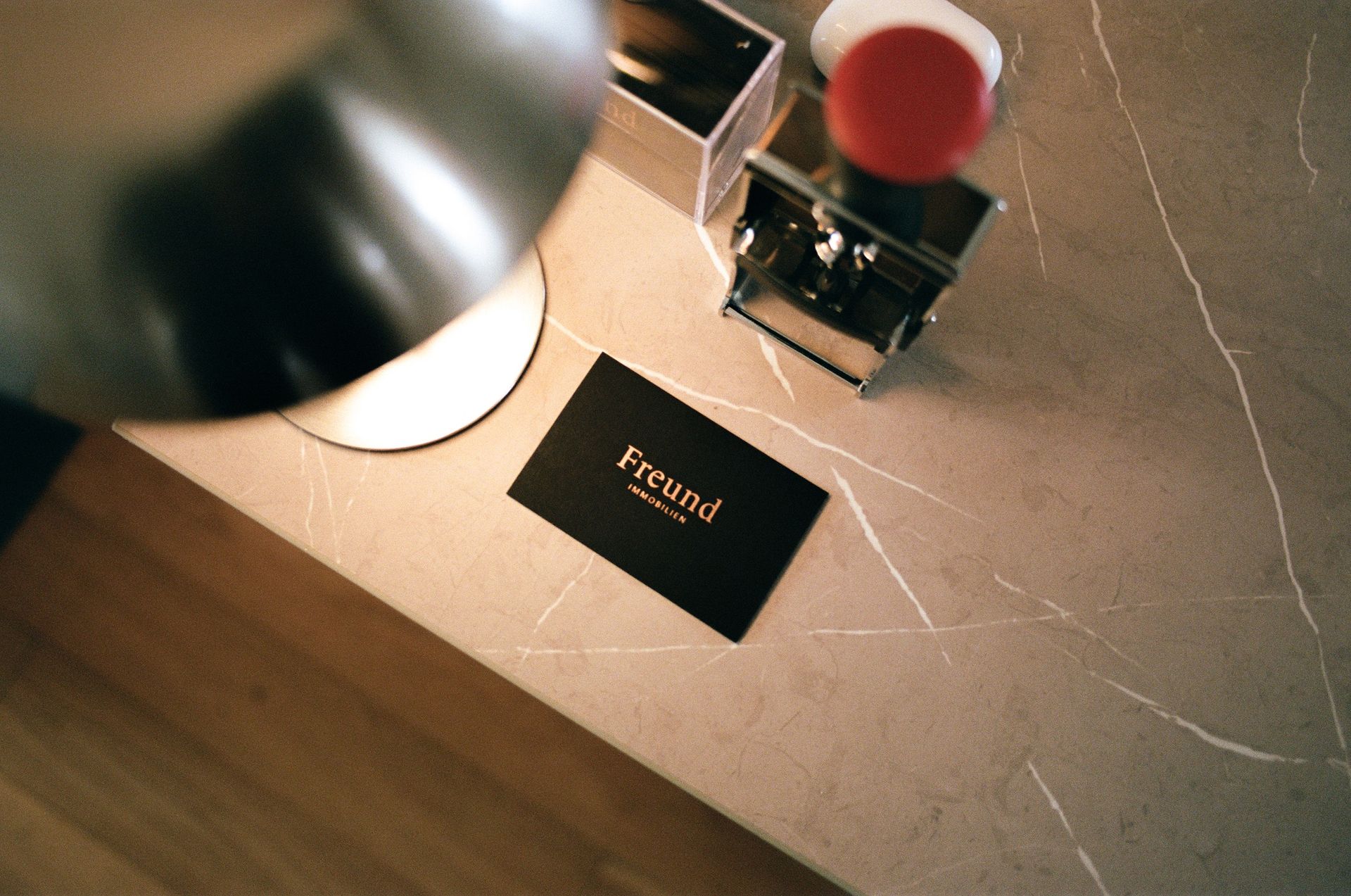 Eine schwarze Visitenkarte mit der Aufschrift "Freund Immobilien" liegt auf einer hellen Marmoroberfläche neben einem teilweise sichtbaren modernen Lampenschirm und einem Parfümflakon mit rotem Deckel.