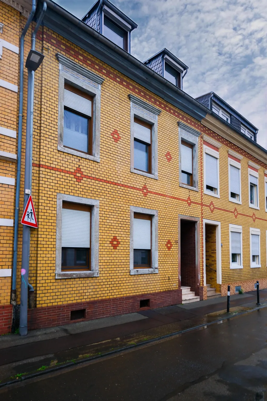 Residential building with yellow and reddish-brown brick pattern, multiple windows with white shutters, and dormer windows under a cloudy sky.