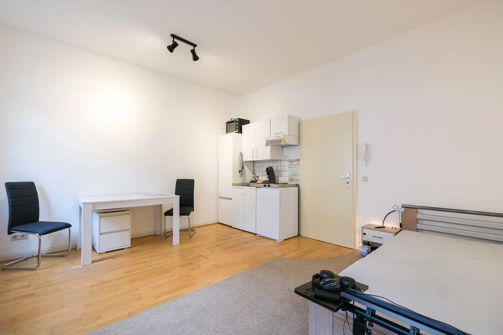 Modern, bright living space with kitchenette, two black chairs, desk, and bed with gray bedding.