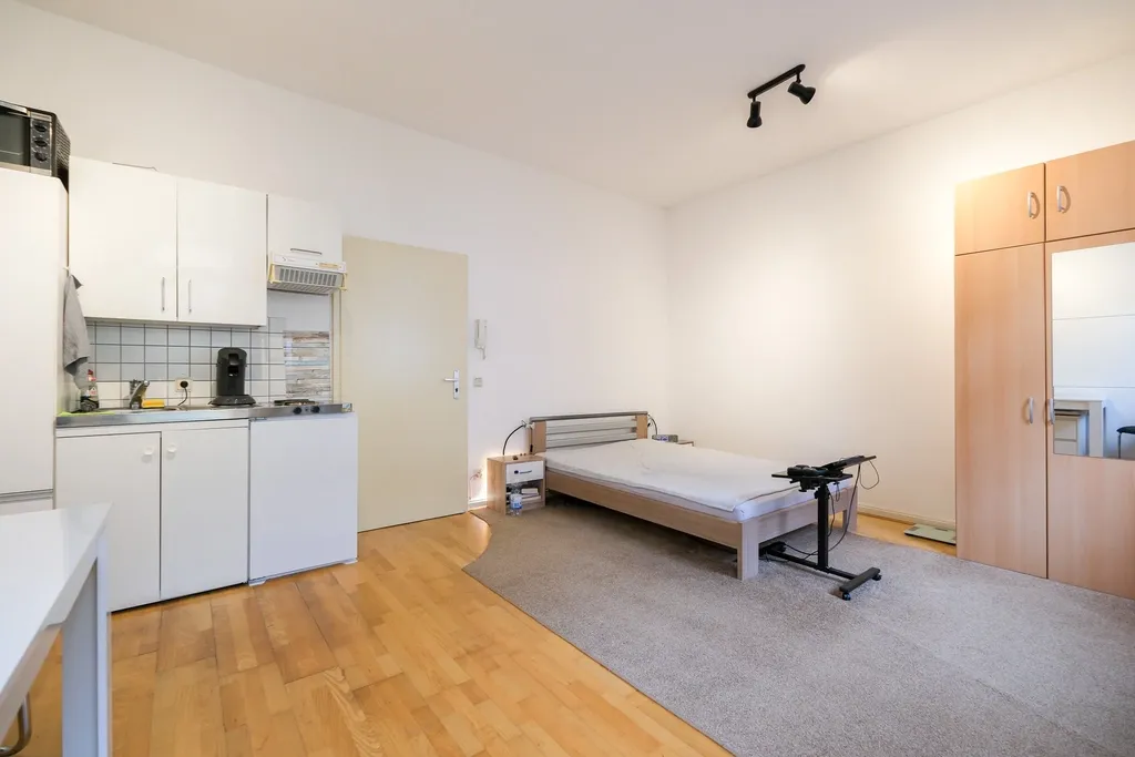 A simply furnished room featuring a single bed on a gray carpet, a small bedside table with a lamp, a kitchenette with white cabinetry, and a large wooden wardrobe with a built-in mirror.