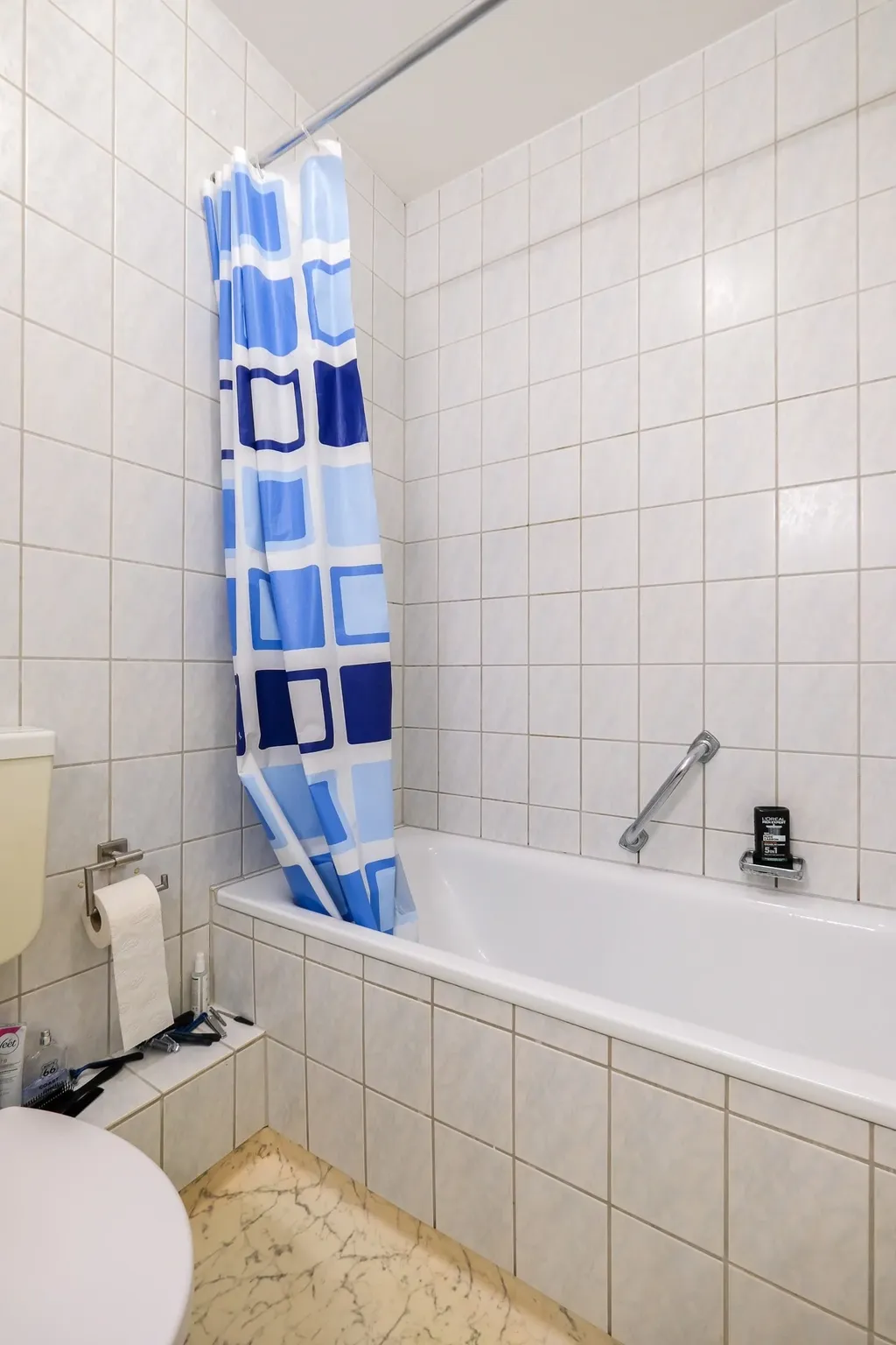 A bathroom with a white tiled bathtub, a blue checkered shower curtain, a toilet paper roll and a toothbrush on the ledge next to the toilet.
