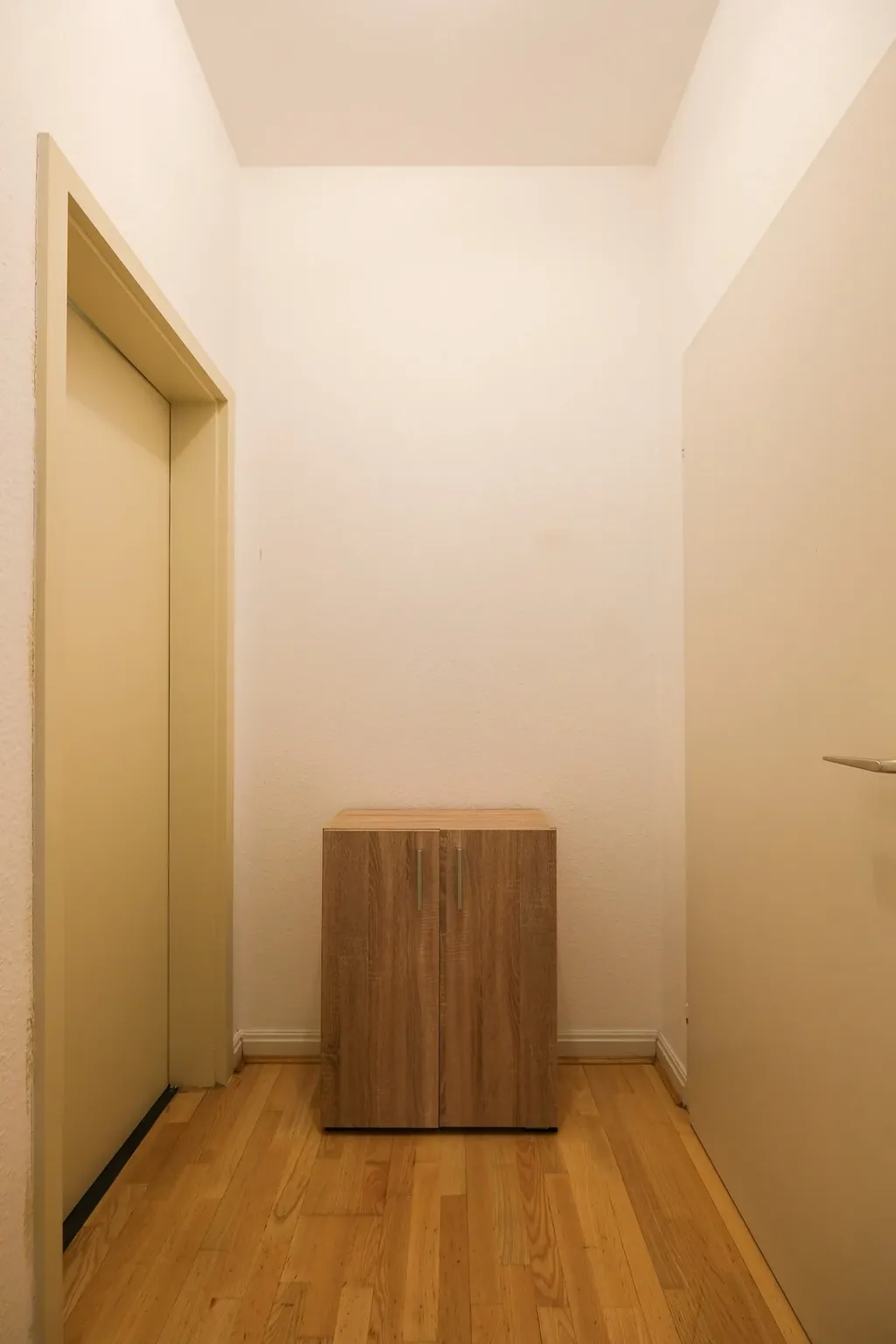 Narrow hallway with wooden floor, a yellow door on the left, a white door with a silver handle on the right, and a small two-door wooden cabinet in the center.