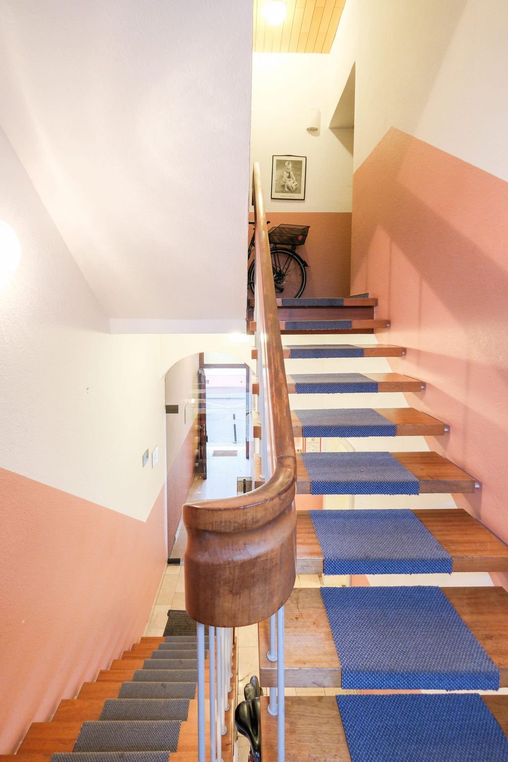 Simple interior stairs with wooden railing, blue carpet runners, and a bicycle at the top of the stairwell in a building with white and pinkish walls.