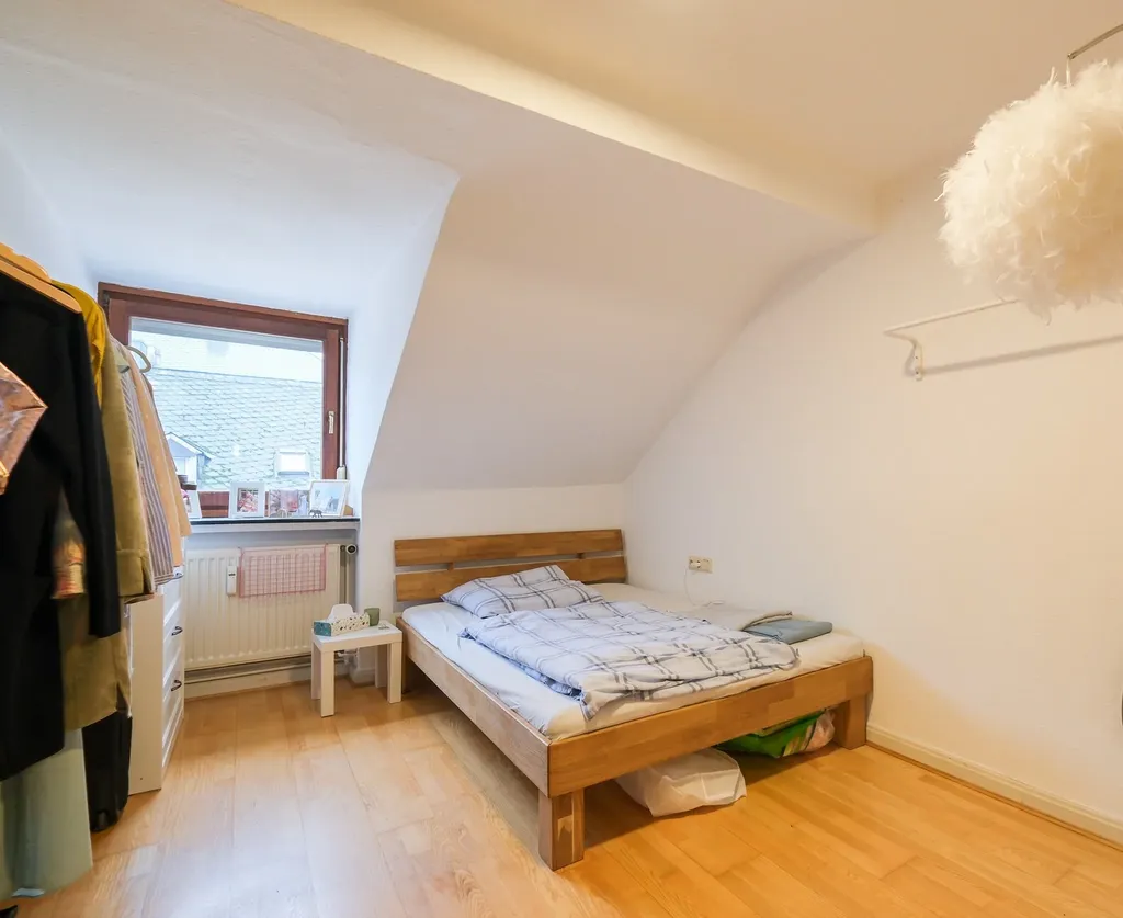 Bedroom with wooden slatted bed, light wooden floor, window overlooking roof tiles, and wardrobe with hanging clothes.
