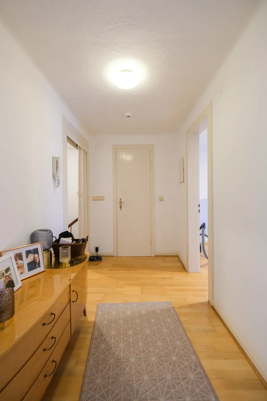 A bright hallway with light wooden flooring, a beige patterned rug in the center, white painted walls, and a closed door at the end. On the left is a dresser with framed family photos and decorative items.