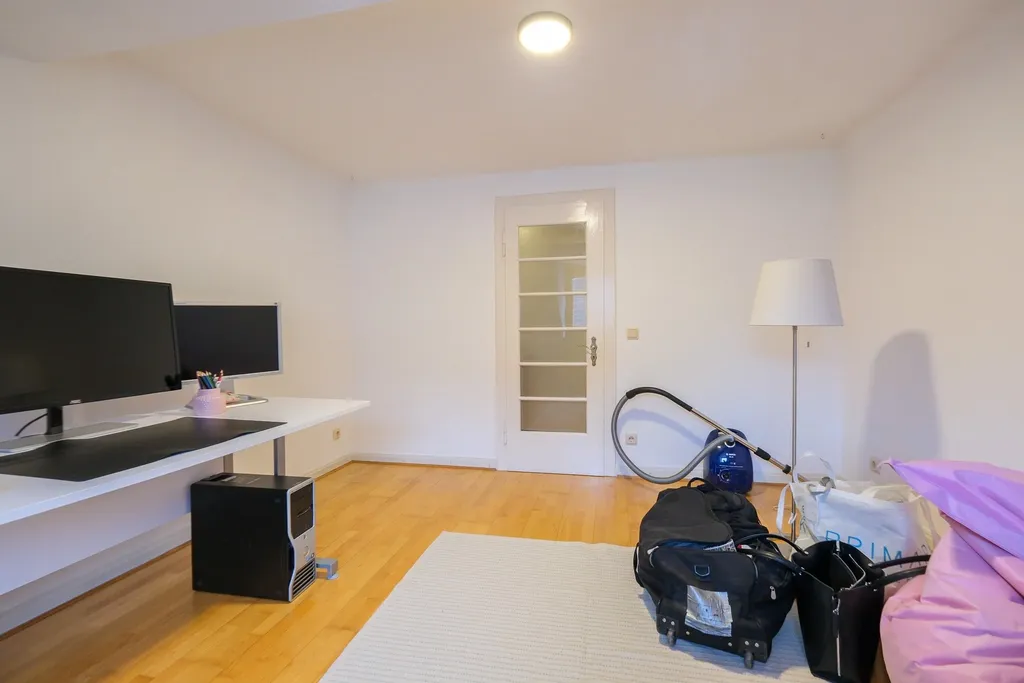 A living room with a white desk and two monitors on the left, a white rug on a wooden floor and a white door in the background, along with a vacuum cleaner, a floor lamp, and suitcases on the right side.