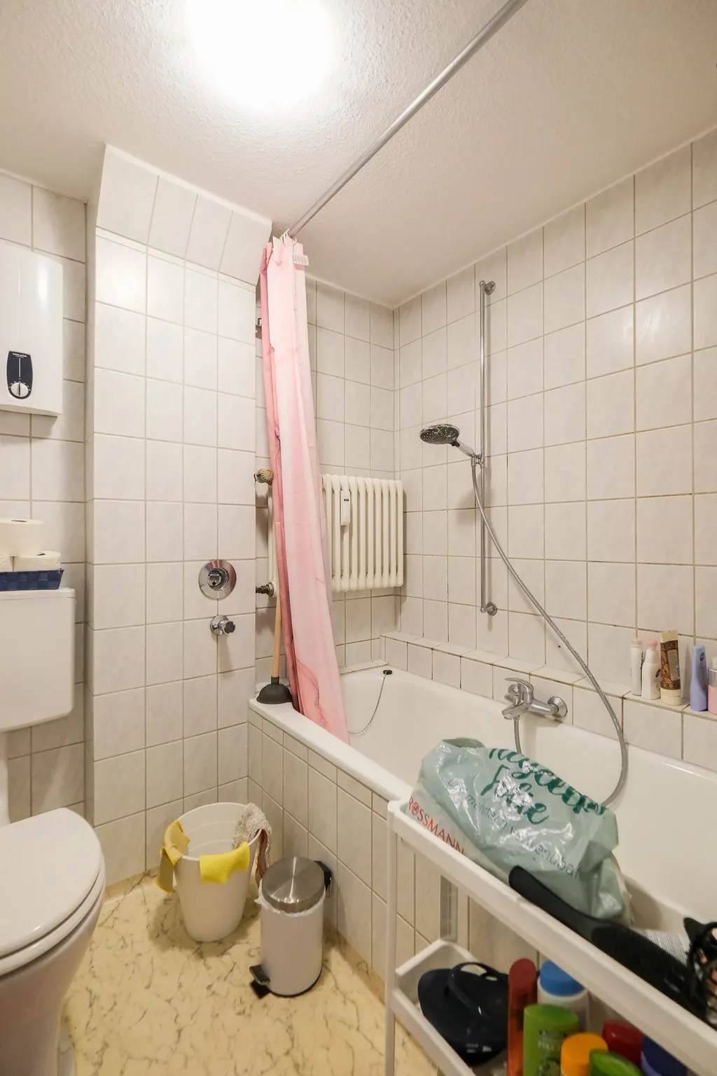 A white tiled bathroom featuring a bathtub with a pink shower curtain, a bucket with yellow gloves, a trash can, and a toilet.