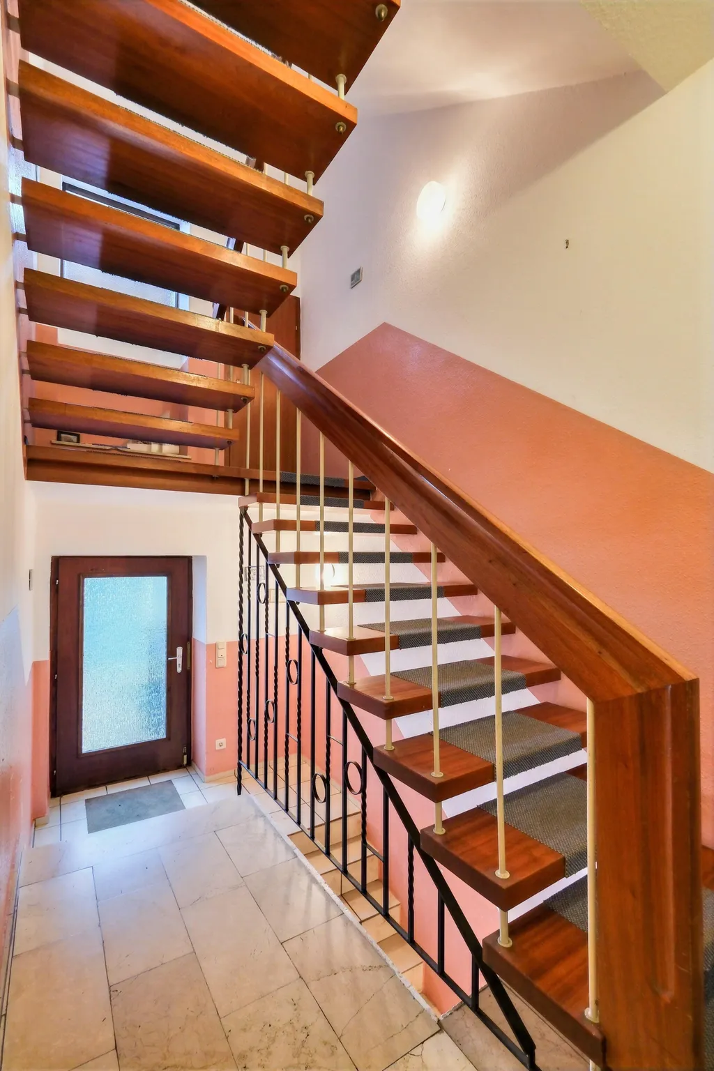 Interior view of a staircase with wooden, semi-floating steps, pink and white painted walls, and a brown door with frosted glass at the bottom.