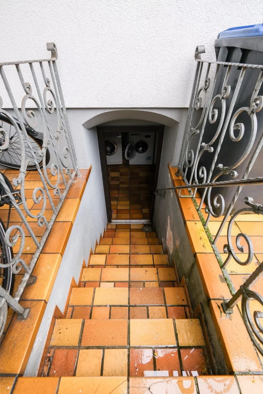 A staircase with orange tiles leads down to a basement room with two washing machines, surrounded by decorative railings and a trash bin.
