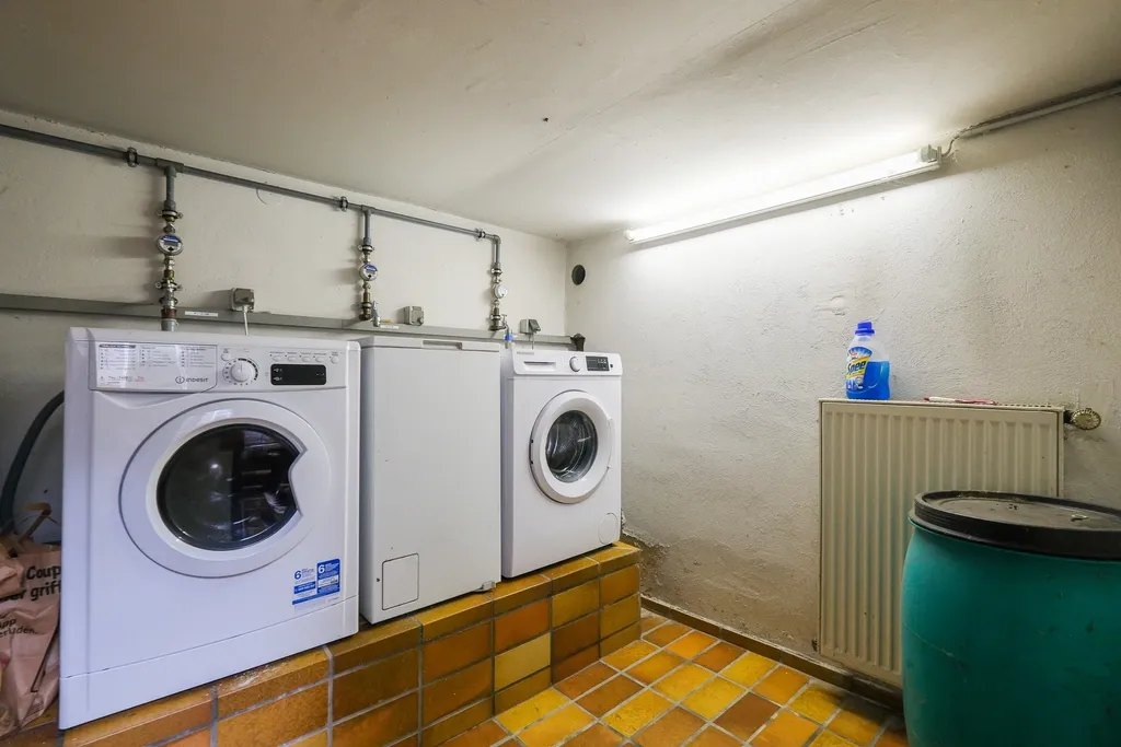A basement room featuring two white washing machines and a refrigerator on a raised tiled platform, next to a radiator with a cleaning bottle on top and a large blue container.