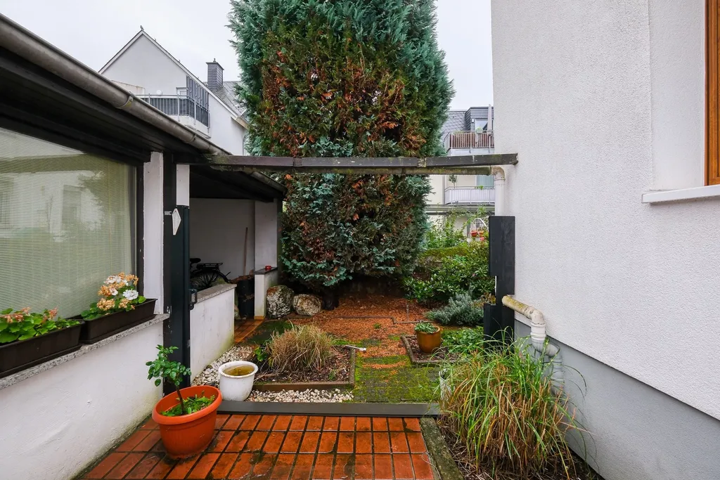 Courtyard with large plants and potted plants between white house walls and a small roof overhang.