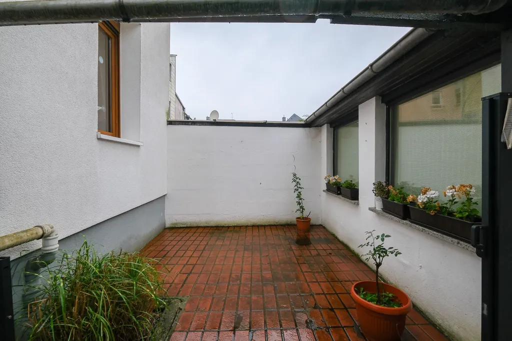 Small enclosed courtyard with red paving, white walls, a few potted plants, and flower boxes on a building window.