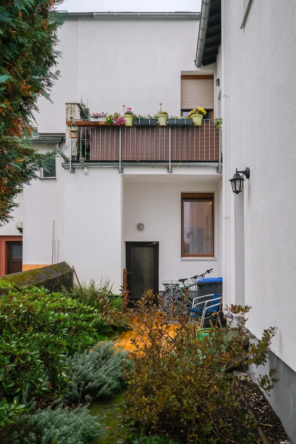 Entrance area of a white multi-family house with a balcony featuring flower pots, surrounded by dense greenery.