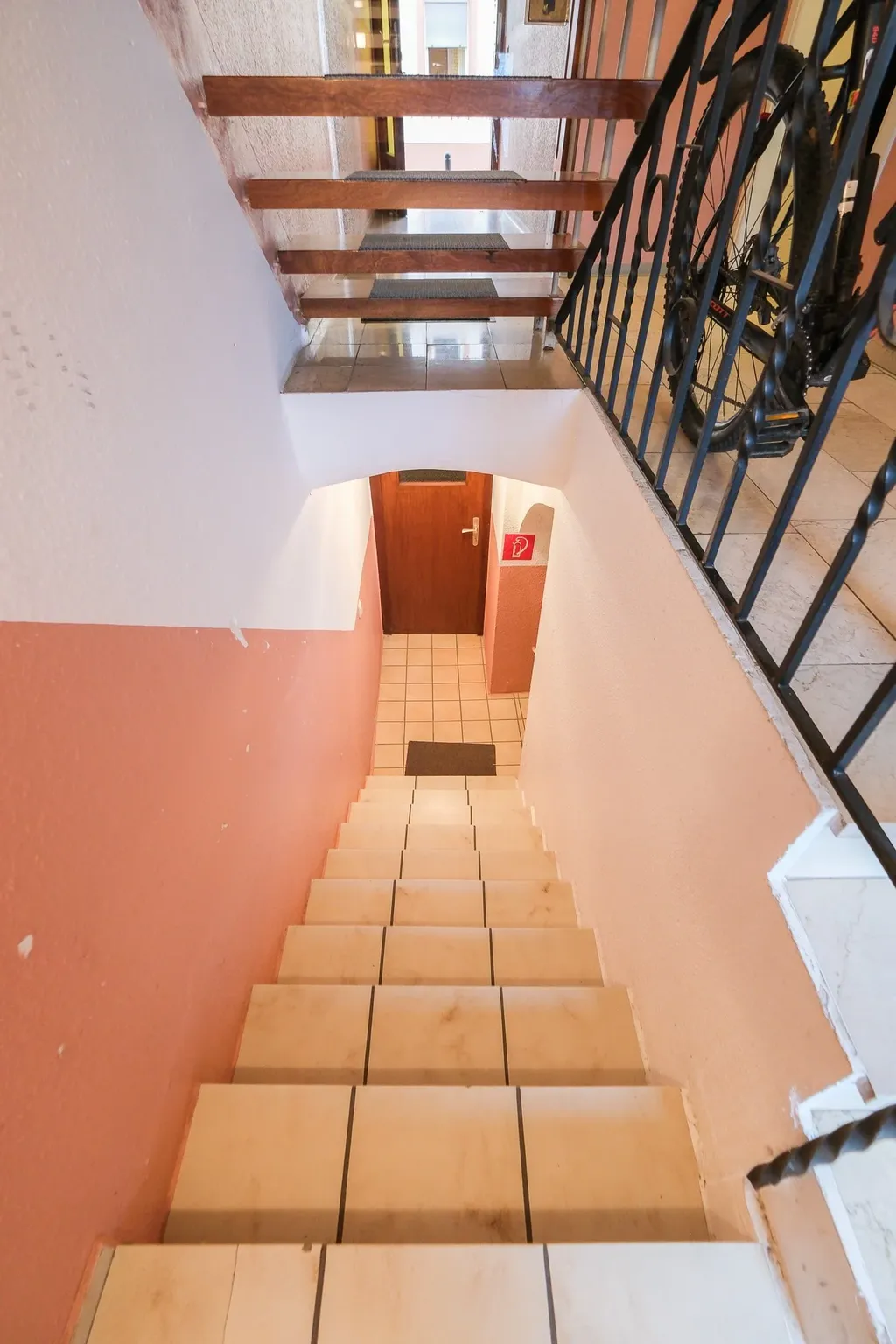 View from above of a stairway with white tiles leading down, surrounded by white and pink walls, with a wooden door at the bottom and a bicycle next to the railing at the top.