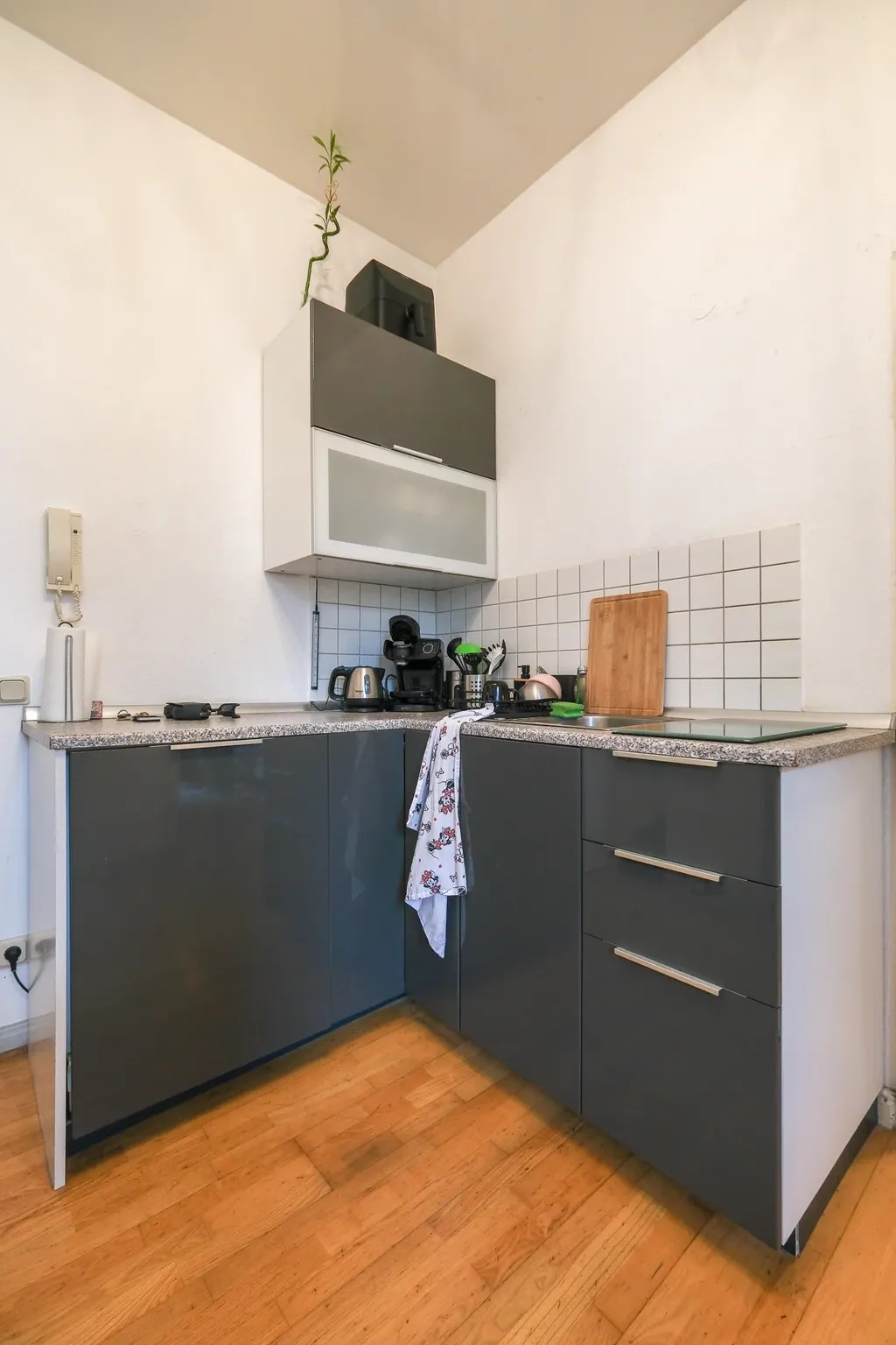 Small modern kitchen with gray cabinets, wooden floor, and white walls, equipped with a coffee machine, toaster, cutting board, and kitchen towel.