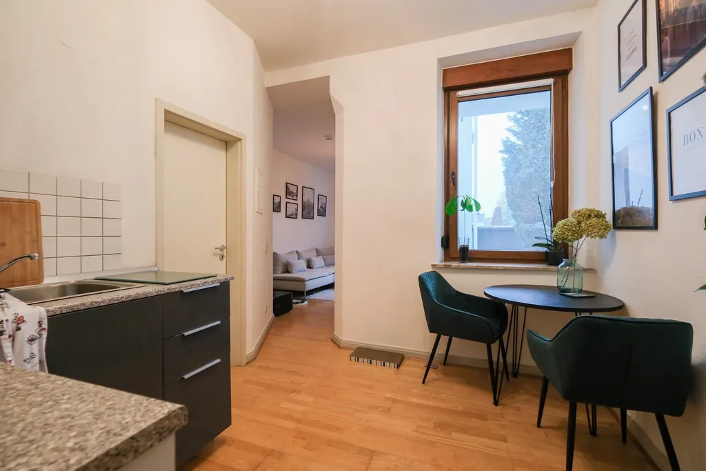 Modern kitchen area with grey cabinets, grey countertop and sink, next to a bright hallway looking into a living room with a sofa and several framed pictures on the wall.