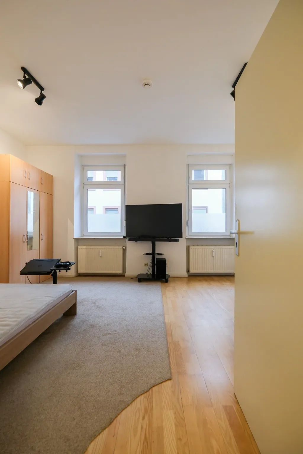 A simply furnished bedroom with a bed without bedding, a large rug, a wardrobe with mirrored doors, and a TV in front of two windows.
