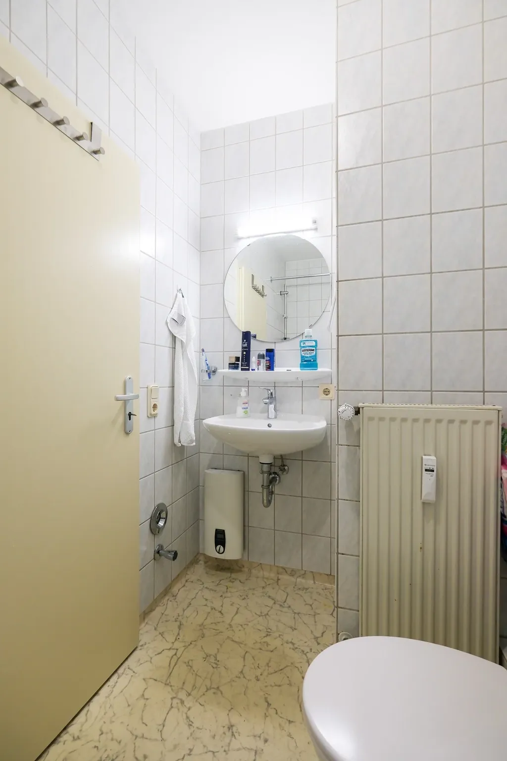 Small bathroom featuring a white sink, round mirror, white tiles, and light brown marble flooring. A towel and various hygiene product bottles are mounted on the wall.