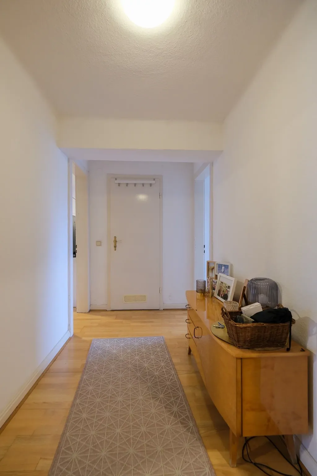 Hallway with light wooden flooring, a patterned runner rug, and a wooden sideboard on the right with picture frames and decorative items, ending at a white door.